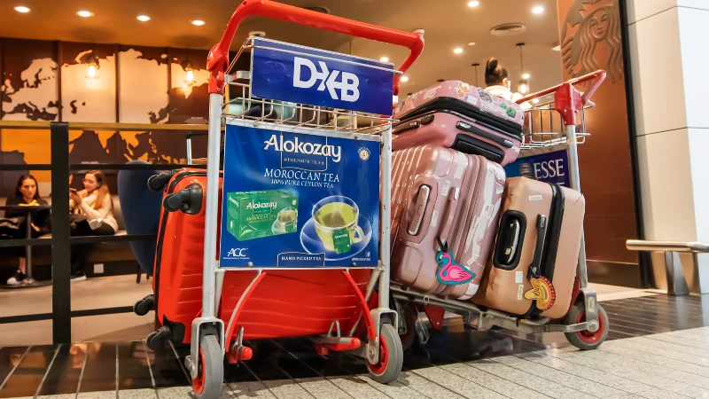 Airport advertising at Dubai International Airport: a DXB trolley advertises Alokozay Moroccan tea beside stacked suitcases.