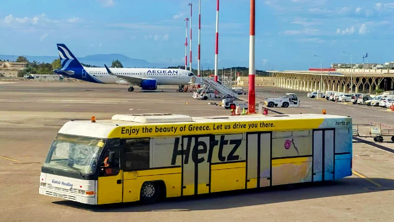 Airport transport advertising in Greece: a yellow Hertz bus reads Enjoy the beauty of Greece. Let us drive you there, on the apron beside an Aegean aircraft.
