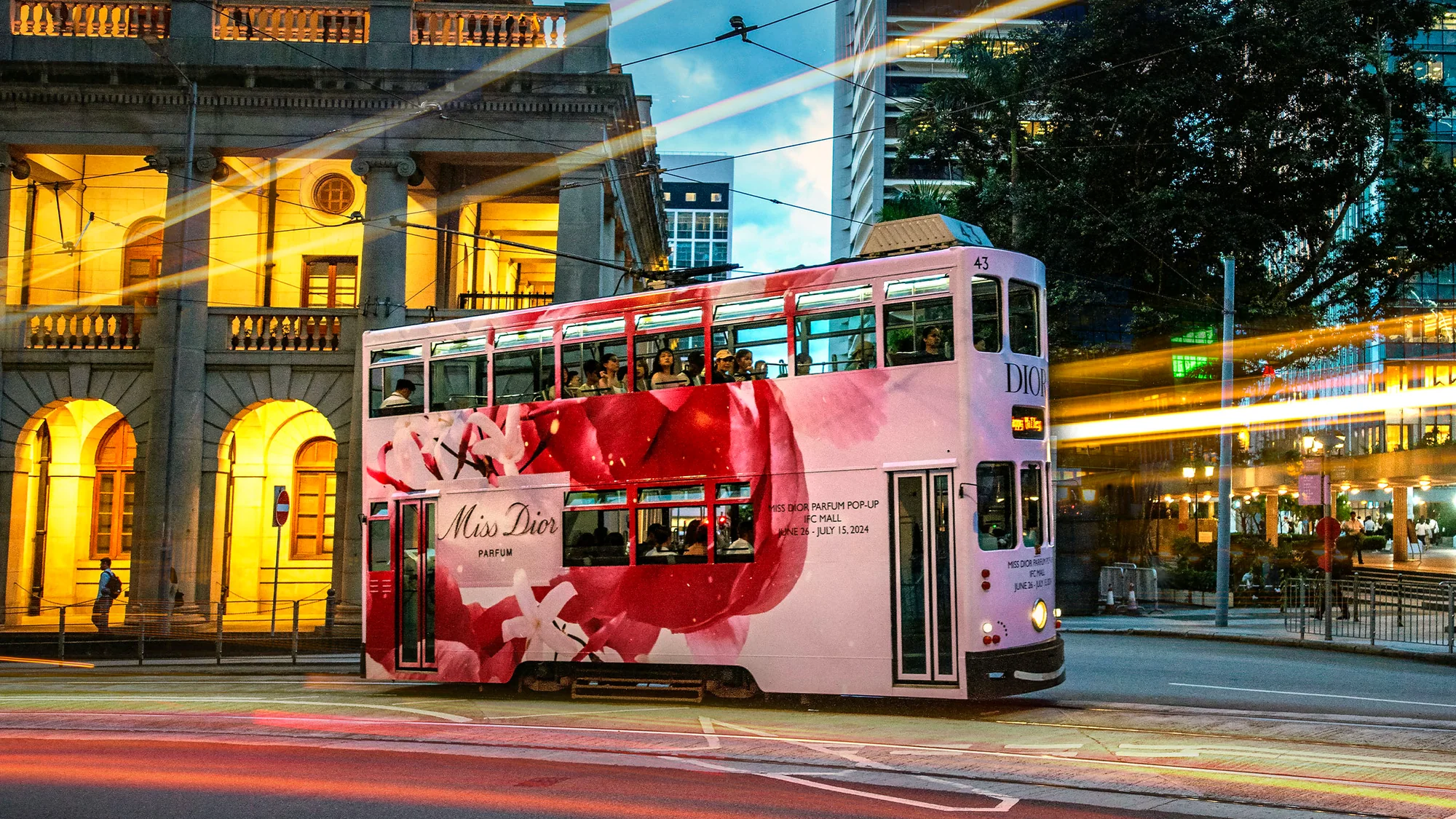 Tram advertising: a double-decker tram wrapped in pink Miss Dior advertising runs past a lit stone building.