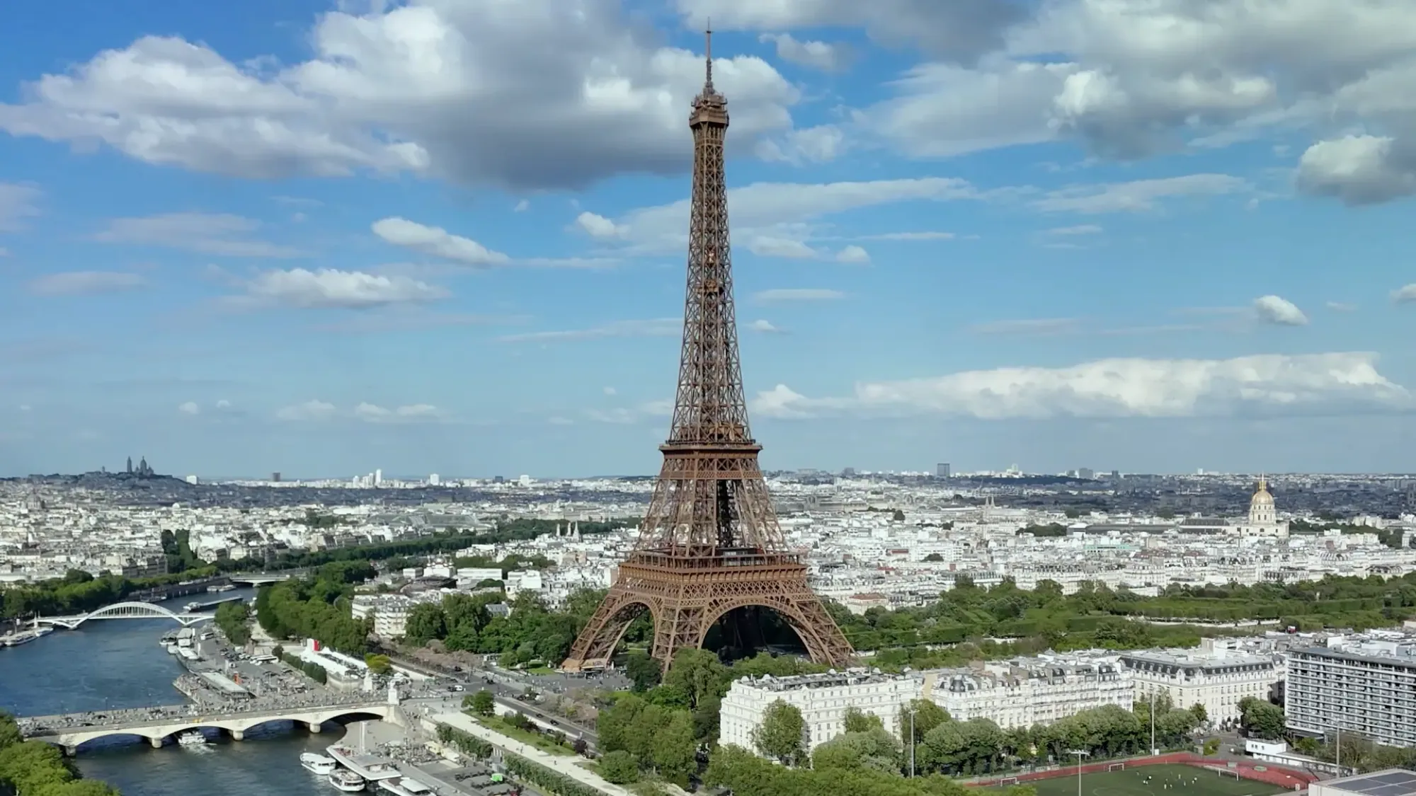 The Eiffel Tower rises above the Seine and the Paris skyline under a cloudy blue sky.
