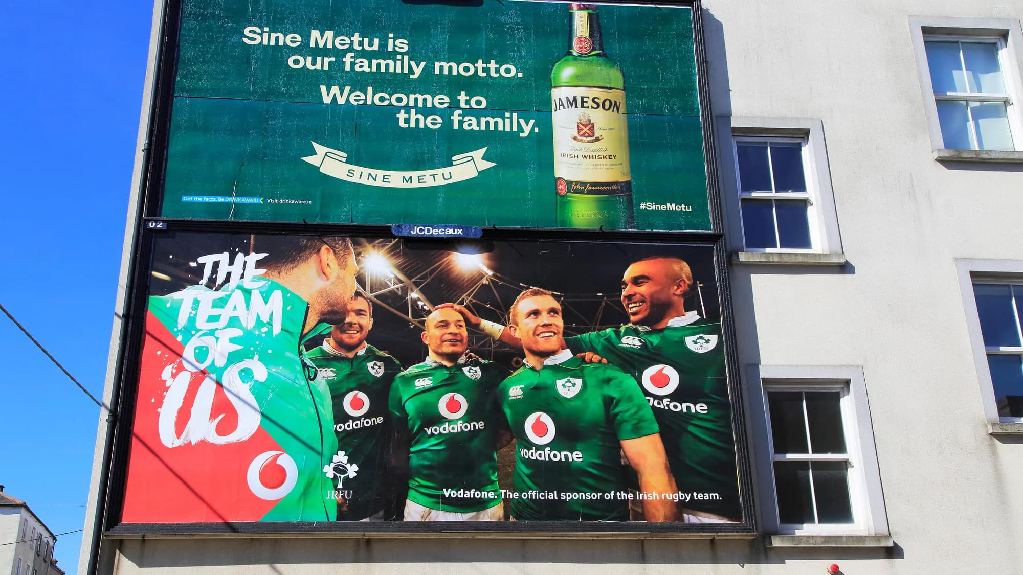 Billboard advertising in Dublin, Ireland: a Jameson board reading Sine Metu sits above a Vodafone poster of the Irish rugby team, The team of us, on a grey building.