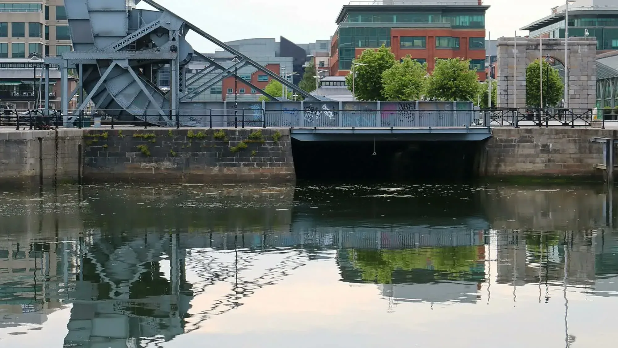 A steel lifting bridge spans a dock, reflected in calm water, with modern offices behind in Dublin.
