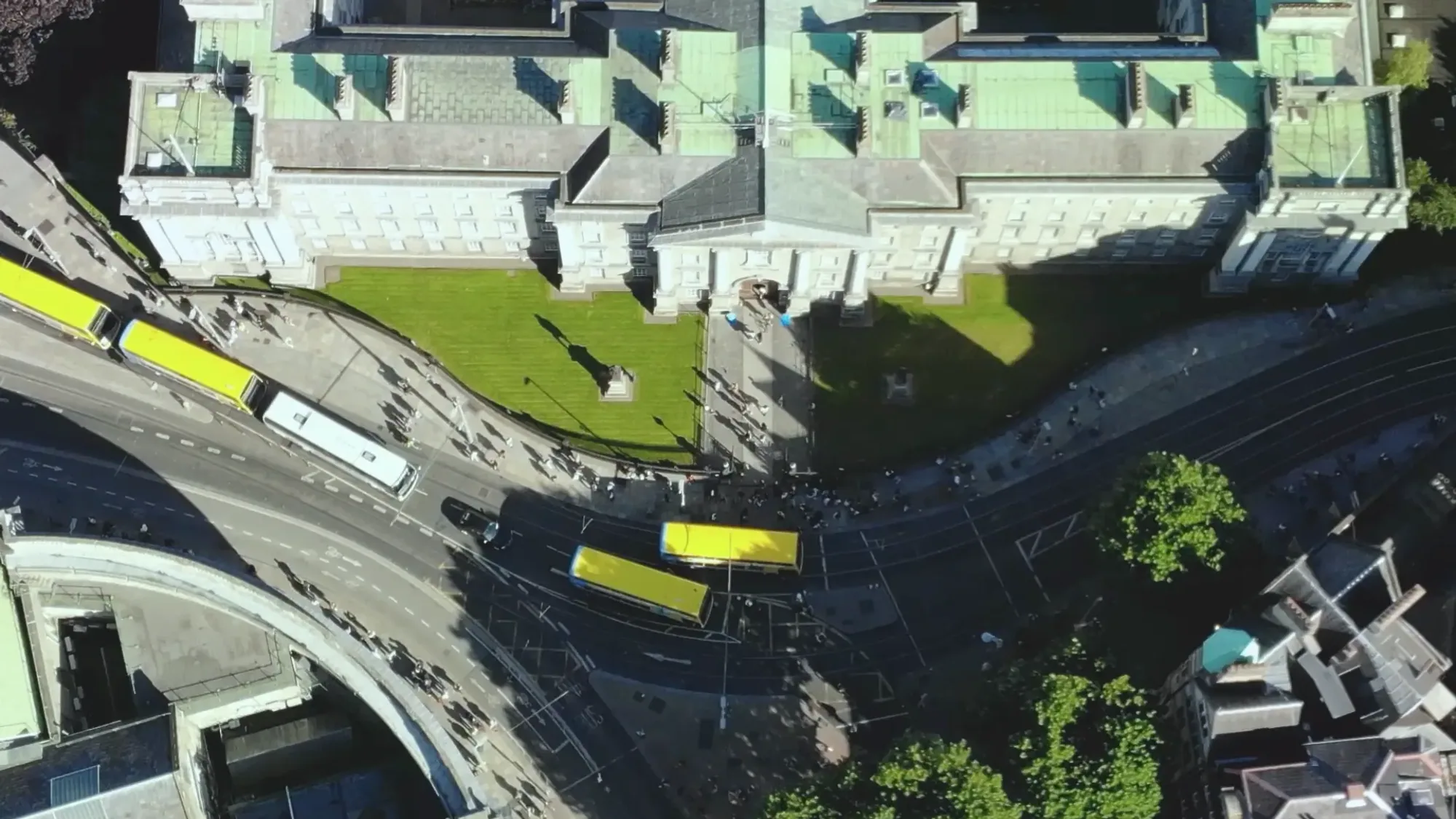 Yellow buses pass a pale stone building with a green copper roof in Ireland, seen from above.