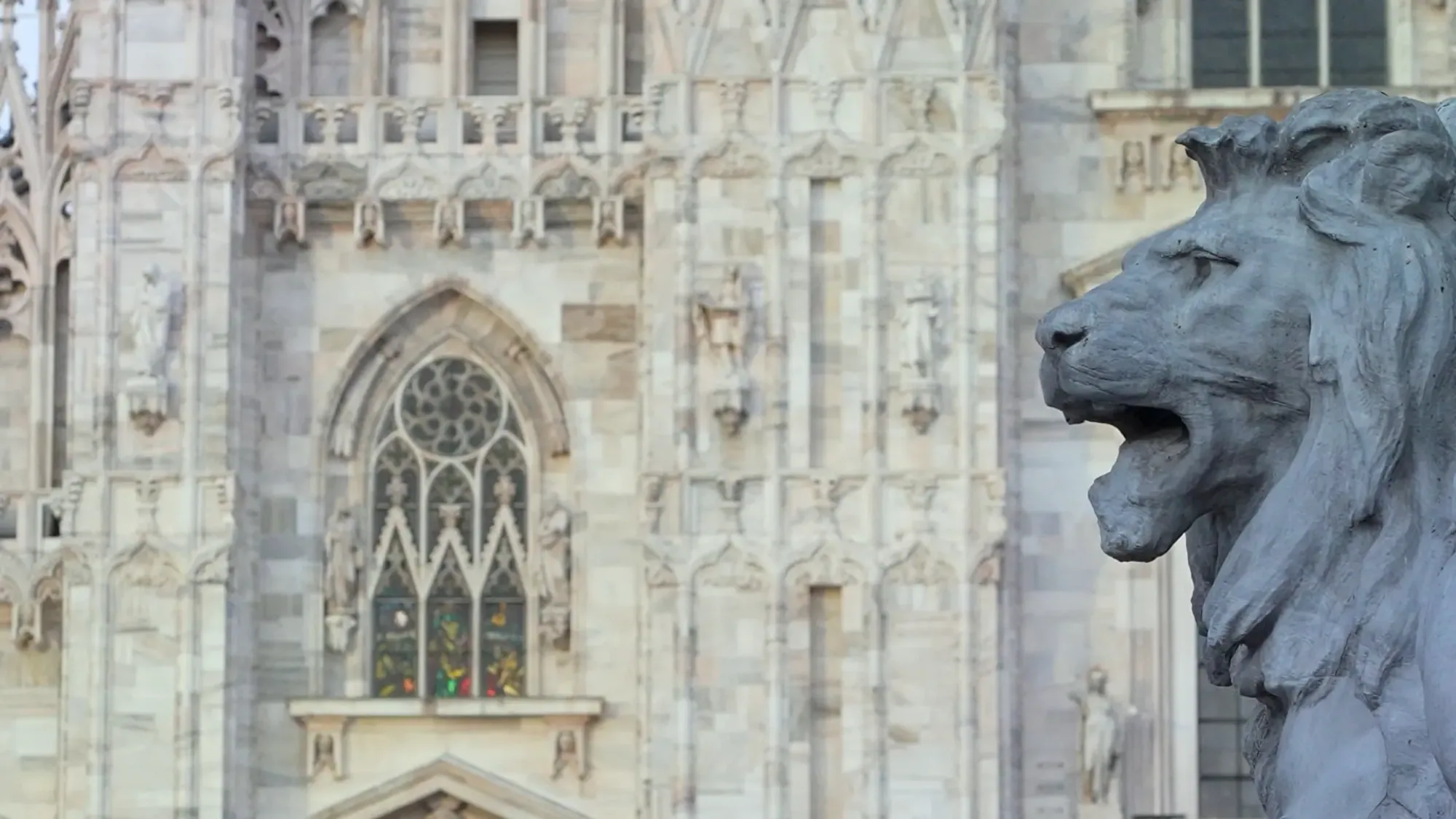 A stone lion stands in front of the carved Gothic facade of Milan Cathedral.