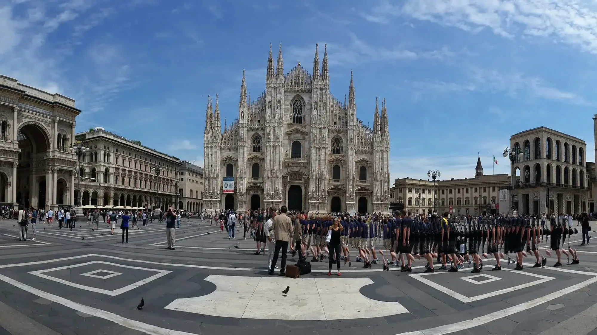 Visitors and a large group in matching outfits gather in the square before Milan Cathedral.