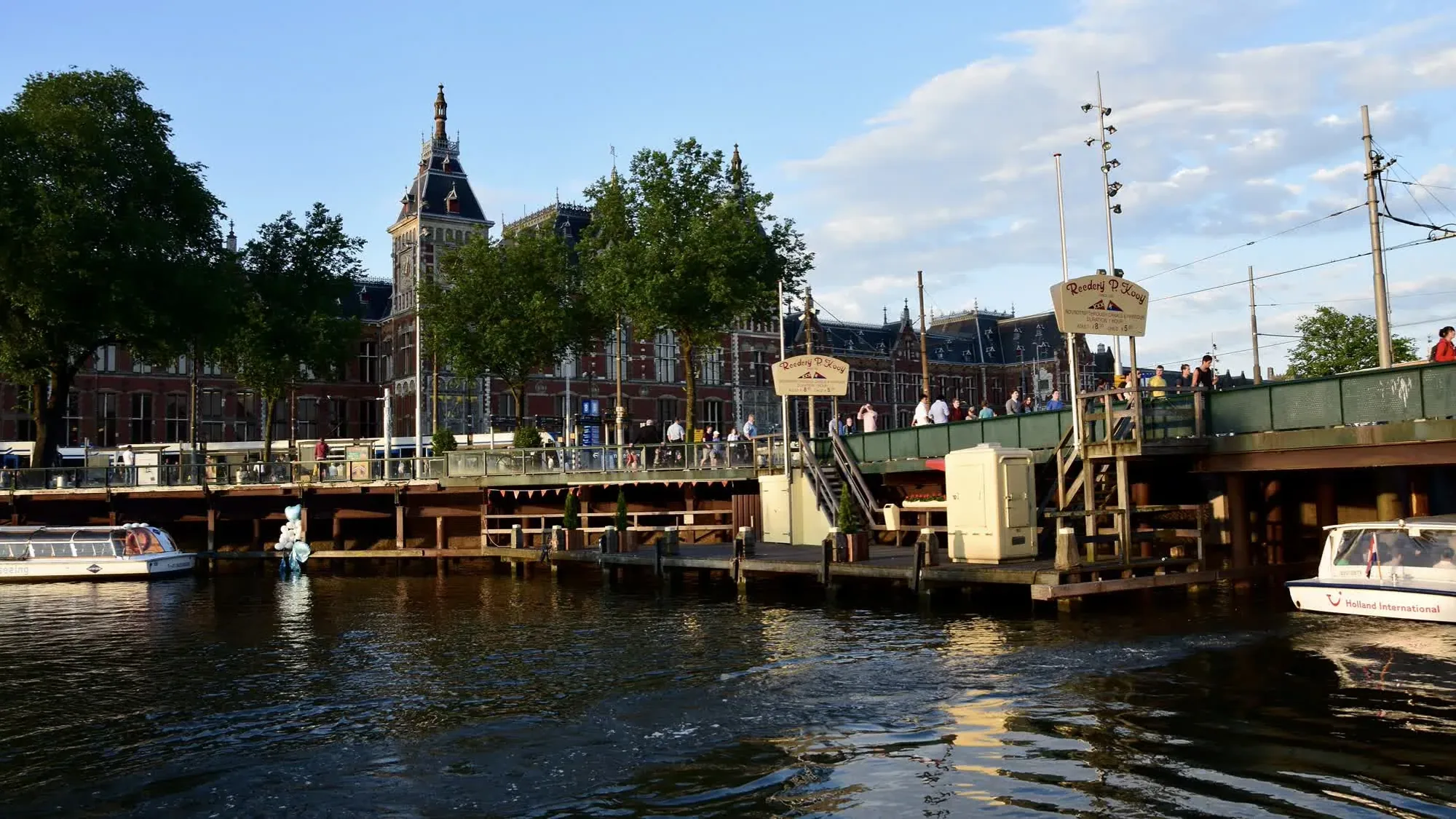 Tour boats pass a canal bridge and the brick towers of Amsterdam Centraal.