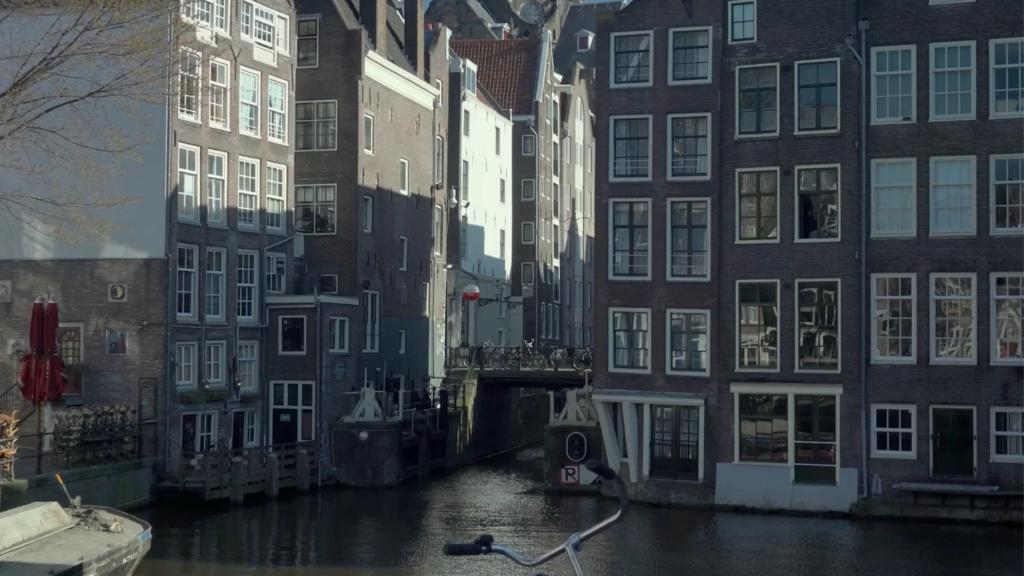 Tall brick canal houses line a narrow waterway, with bicycles parked on a bridge in the Netherlands.