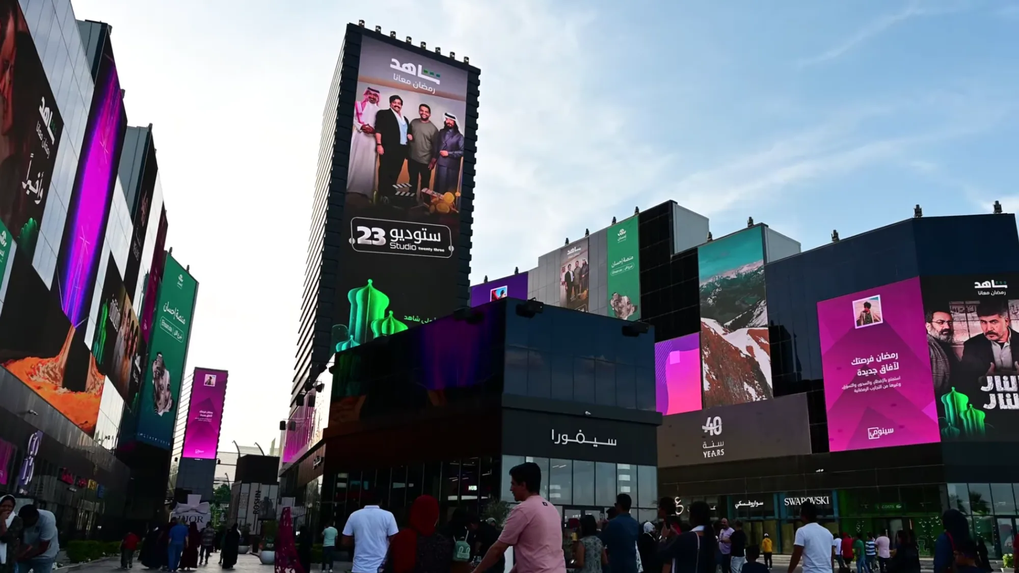Digital billboard advertising in Saudi Arabia: pedestrians pass tall screens and building wraps carrying Arabic advertising.