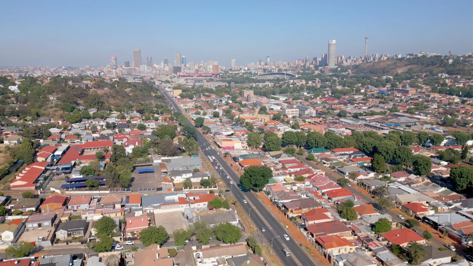 An aerial view shows red-roofed suburbs and a highway leading towards a high-rise skyline in South Africa.