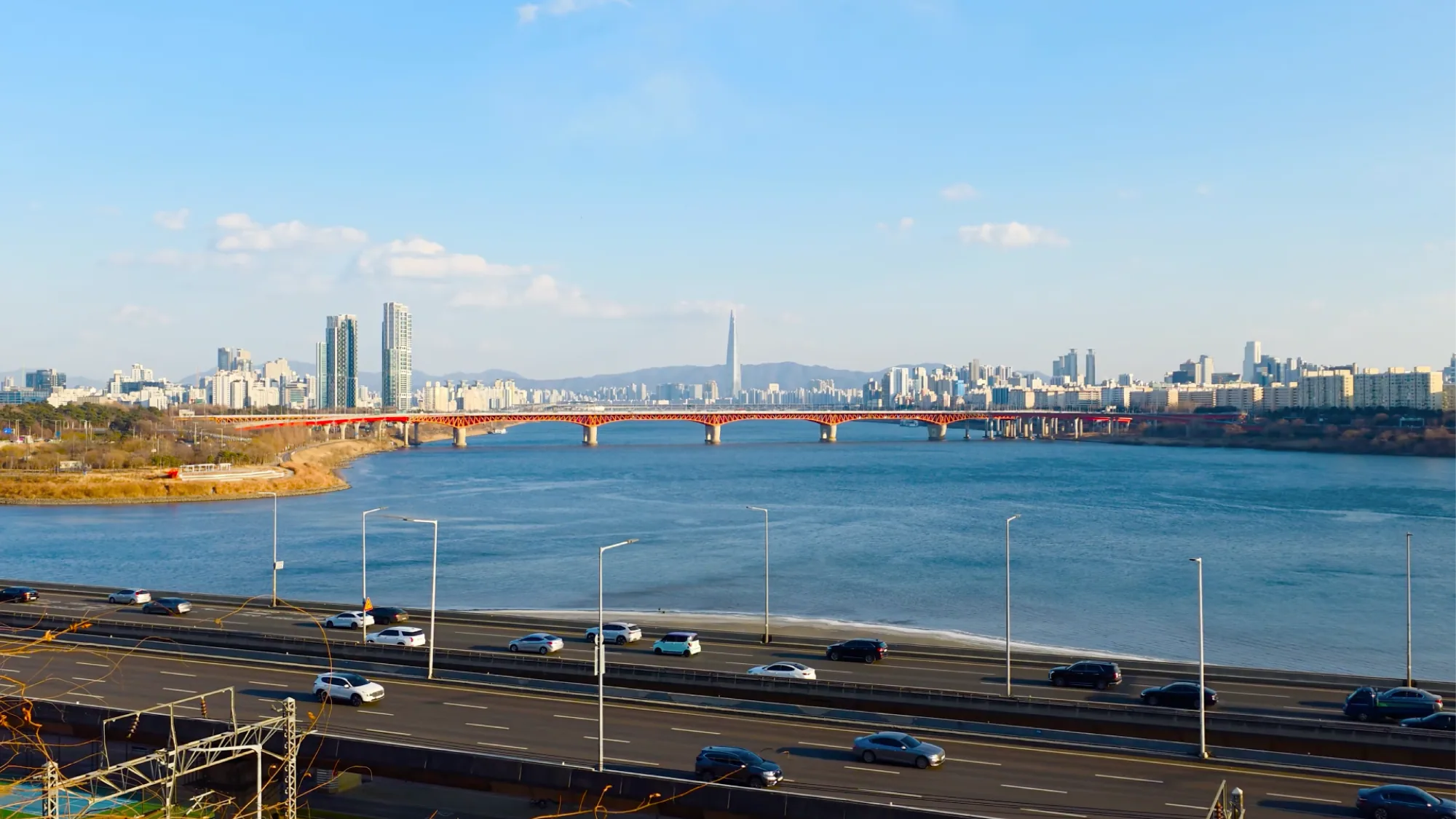 A red bridge crosses a wide river towards the Seoul skyline, with highway traffic in front.
