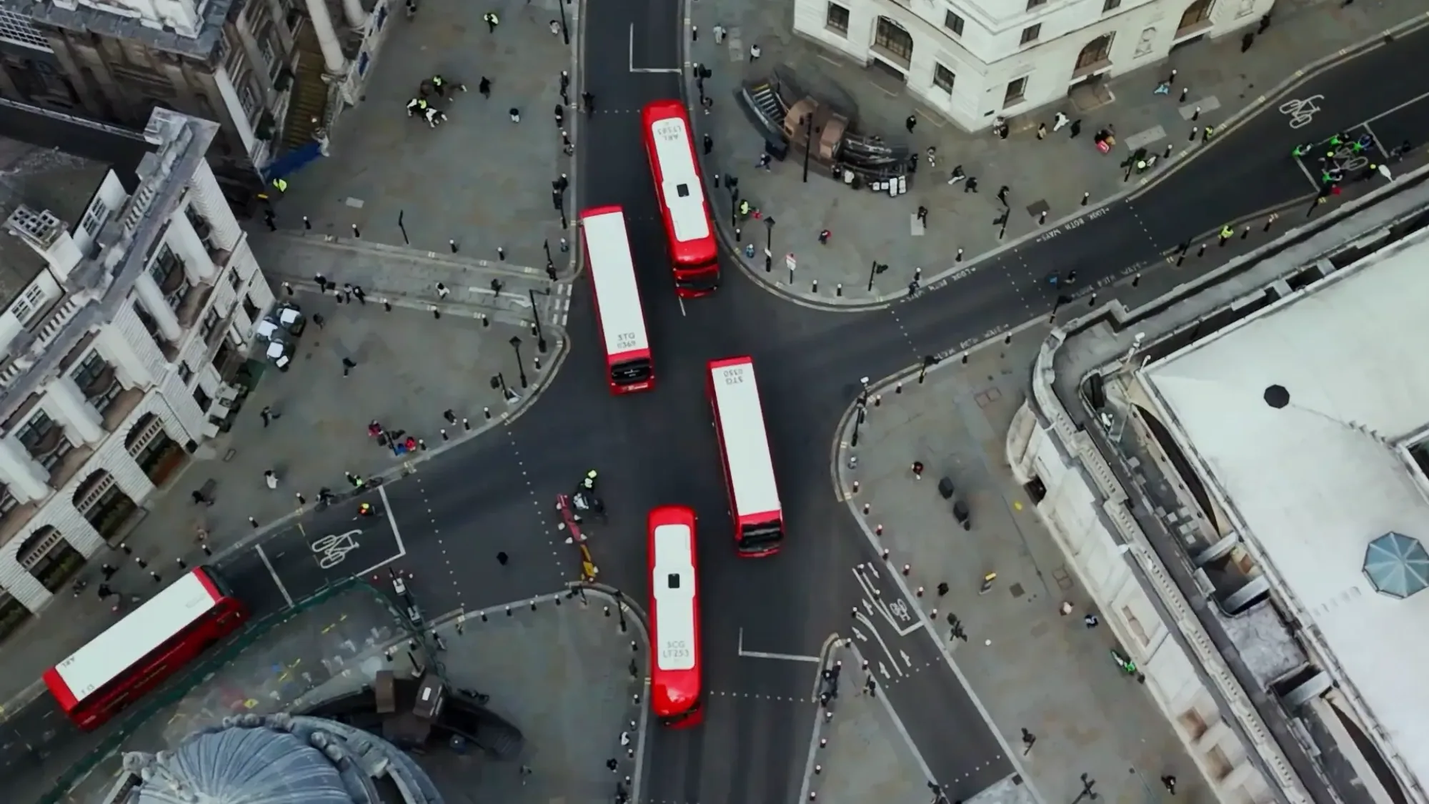 Red buses cross a junction among stone buildings in London, seen from above.