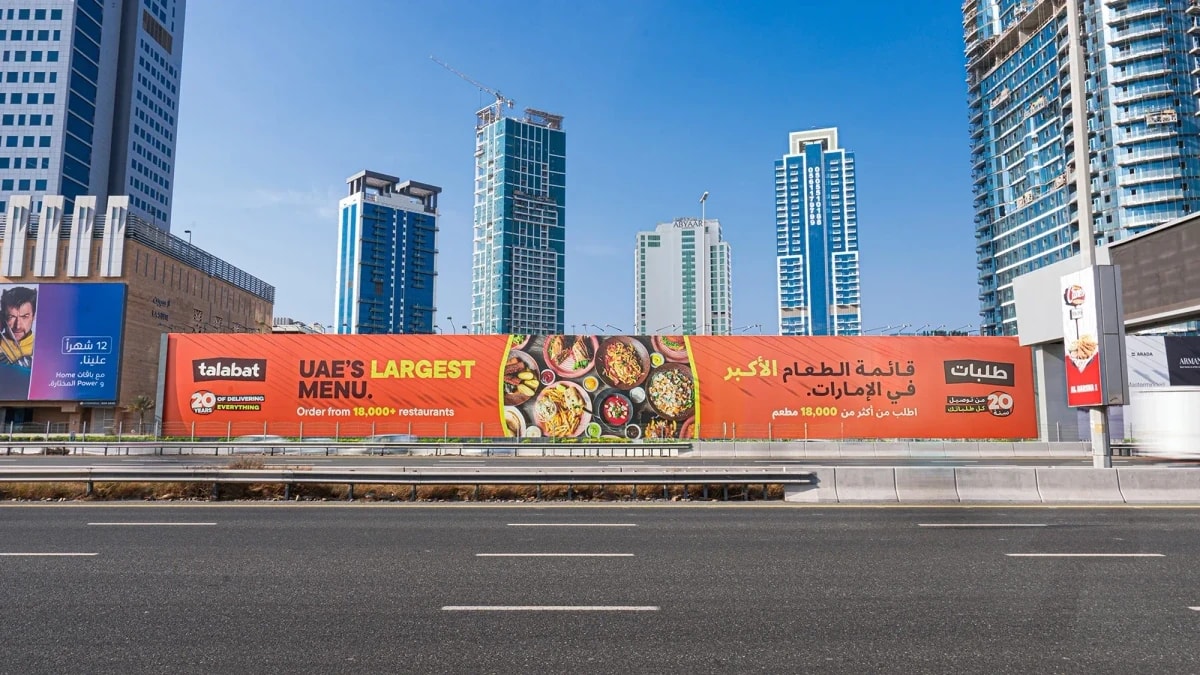 Hoarding advertising in the UAE: a long roadside Talabat board reads UAE's largest menu and Order from 18,000+ restaurants, with glass towers behind it.