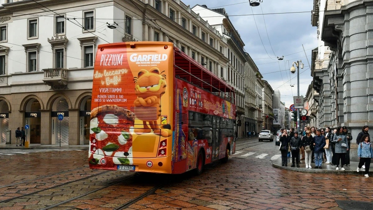 Bus rear advertising: the back of an orange double-decker carries a Pizzium advert with Garfield and a pizza, on a cobbled street with tram tracks, past a Fendi store.