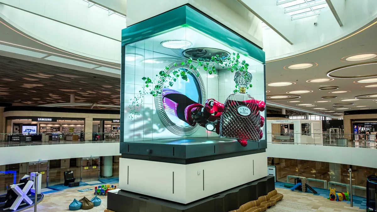 Digital out-of-home advertising in a mall atrium: a cube screen shows a perfume bottle bursting through a circular frame.
