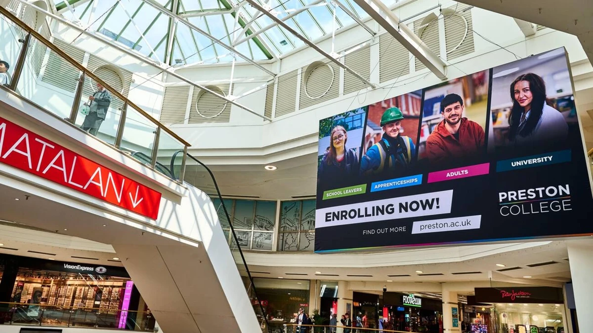 Mall digital screen advertising: a hanging screen beside a Matalan sign shows four portraits for Preston College, with the line Enrolling now and preston.ac.uk.