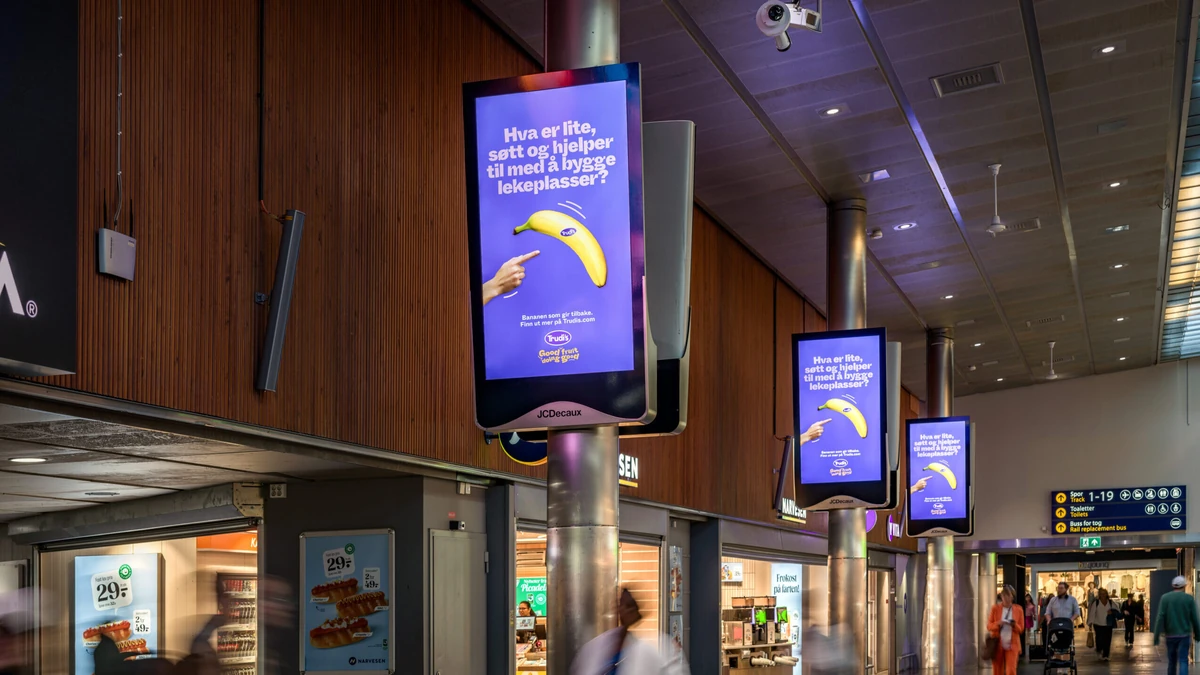Programmatic screen advertising: JCDecaux column screens in a concourse show a purple banana advert in Norwegian, a hand pointing at the fruit, beside shops and a departures board.