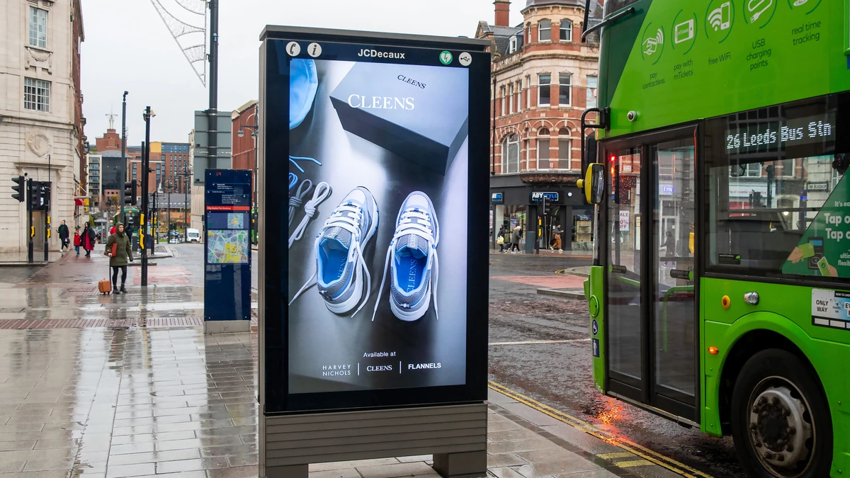 Guaranteed programmatic advertising: a JCDecaux street screen on a wet pavement shows Cleens trainers, available at Harvey Nichols and Flannels, beside a green bus.