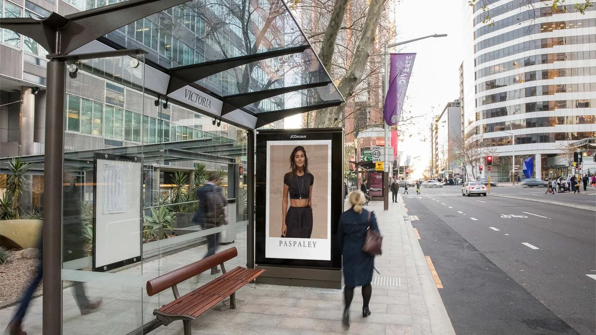 Street furniture advertising: a glass bus shelter on a city pavement carries a Paspaley poster of a woman.