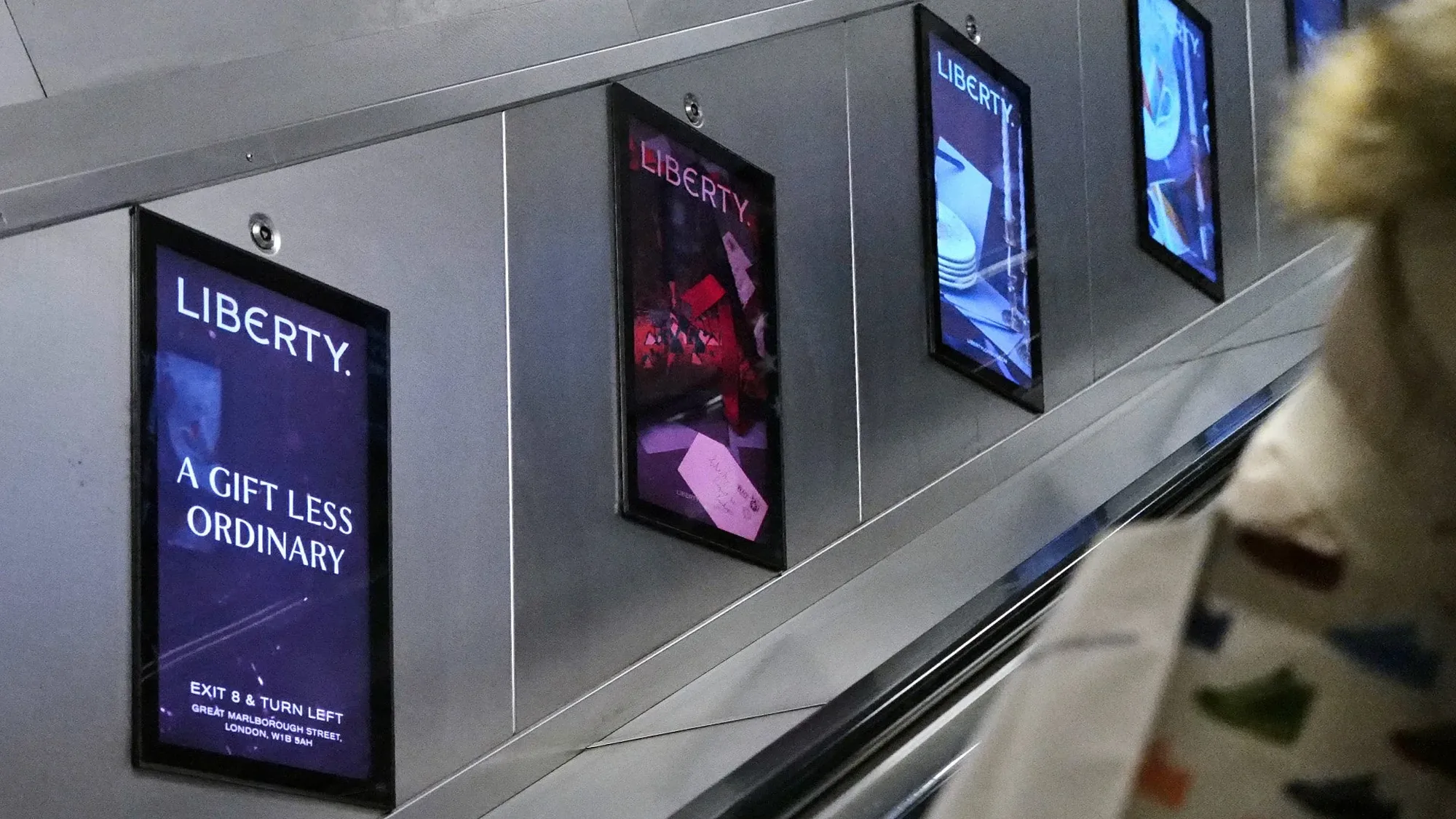 London Underground advertising in the United Kingdom: Liberty posters reading A gift less ordinary line the wall beside an escalator.