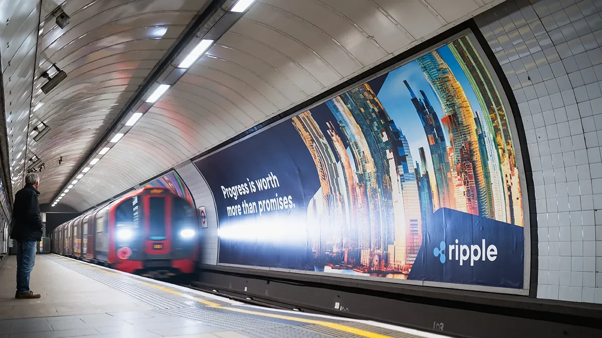 London Underground platform advertising: a long Ripple poster along the tunnel wall reads Progress is worth more than promises, as a red train arrives.