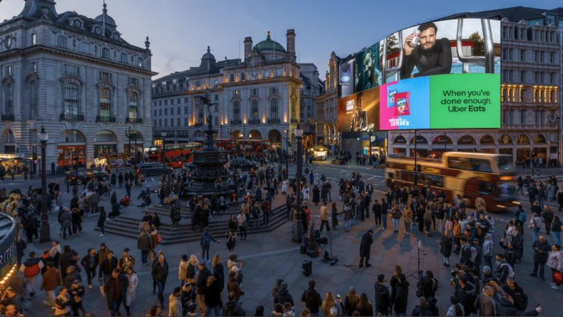 Digital billboard advertising at Piccadilly Circus in London, in the United Kingdom: curved screens show Diet Coke, Uber Eats and Doritos above the Eros statue, a crowd and a red bus at dusk.
