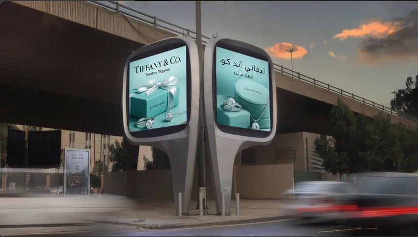 Digital out-of-home advertising in Saudi Arabia: twin rounded roadside screens show Tiffany & Co. jewellery ads in English and Arabic beside a highway overpass at dusk.